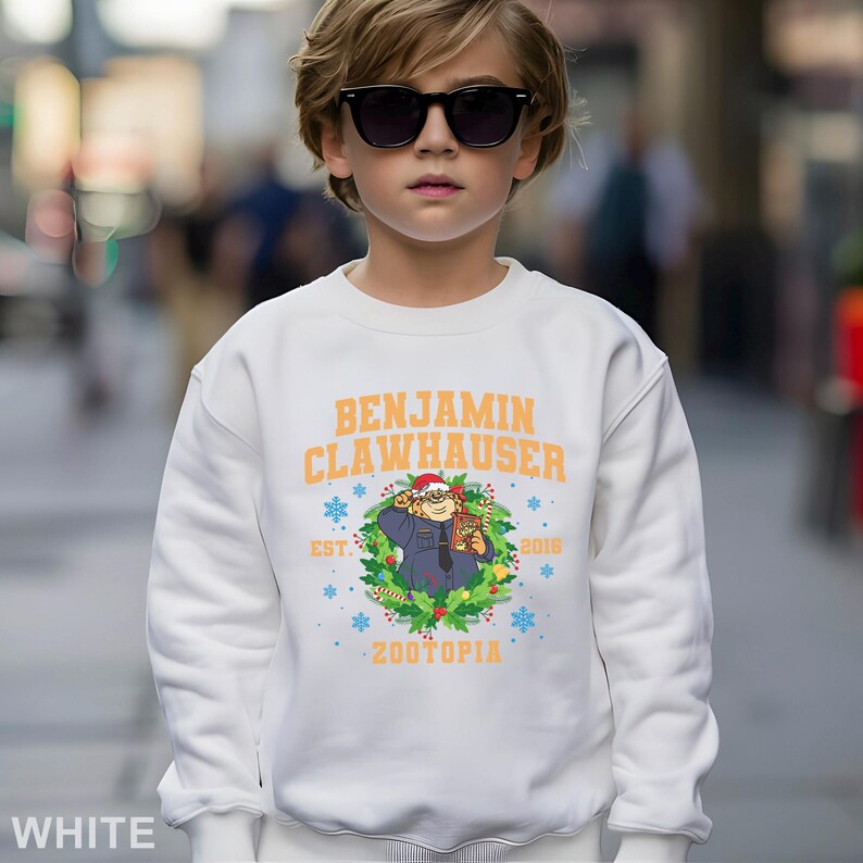 Pu&ograve; includere: Felpa bianca con la scritta "BENJAMIN CLAWHAUSER" in arancione e un'illustrazione di un personaggio di Zootopia. Il design include fiocchi di neve, l'anno 2016 e la parola "ZOOTOPIA".