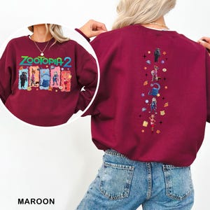 K&ouml;nnte beinhalten: Burgunderfarbenes Sweatshirt mit dem Text "ZOOTOPIA 2" in Gr&uuml;n, Orange und Blau. Die Grafik auf der Vorderseite zeigt Charaktere aus dem Film. Die R&uuml;ckseite des Sweatshirts hat ein vertikales Design mit weiteren Charakteren und Sternen.