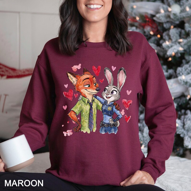 Pu&ograve; includere: Felpa bordeaux con un'illustrazione ad acquerello di Nick Wilde e Judy Hopps di Zootropolis, circondati da cuori rossi. I personaggi si abbracciano. La parola "MAROON" &egrave; stampata in basso.