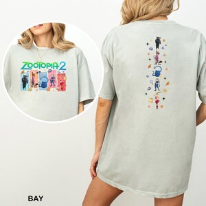Op de afbeelding: Lichtgrijze t-shirt met de tekst "ZOOTOPIA 2" en kleurrijke karakterillustraties op de voorkant. De achterkant van de shirt heeft verticale illustraties van personages en objecten uit de film.