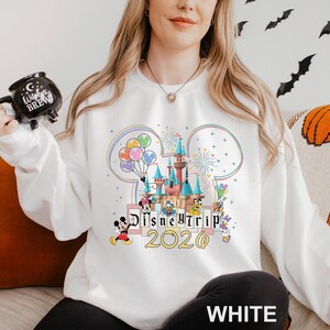 Puede incluir: Sudadera blanca con un dise&ntilde;o de castillo de Disney, orejas de Mickey Mouse, globos y fuegos artificiales. El texto "Disney Trip 2020" est&aacute; debajo. La sudadera es de estilo casual.