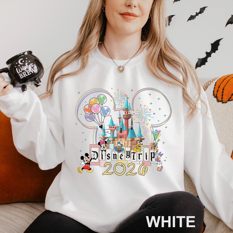Puede incluir: Sudadera blanca con un dise&ntilde;o de castillo de Disney, orejas de Mickey Mouse, globos y fuegos artificiales. El texto "Disney Trip 2020" est&aacute; debajo. La sudadera es de estilo casual.