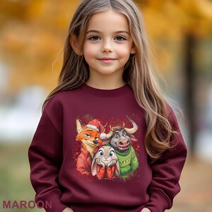 May include: A maroon sweatshirt featuring a colorful graphic of cartoon characters, including a fox wearing a Santa hat, a bull, and a bunny. The word "MAROON" is printed at the bottom of the sweatshirt. The sweatshirt is worn by a young girl.