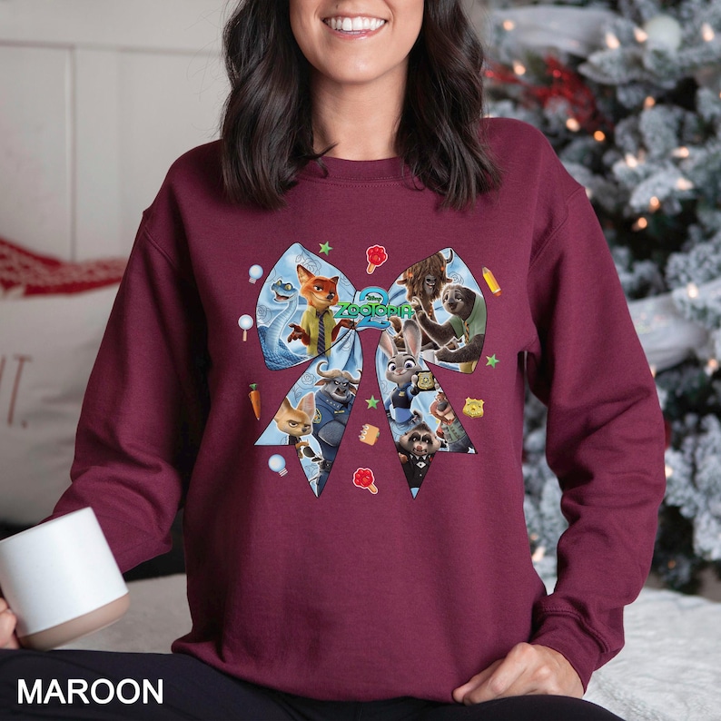 May include: Maroon sweatshirt featuring a large bow design with characters from the movie Zootopia. The bow is light blue with images of the characters and the movie title. The sweatshirt is a solid maroon color.