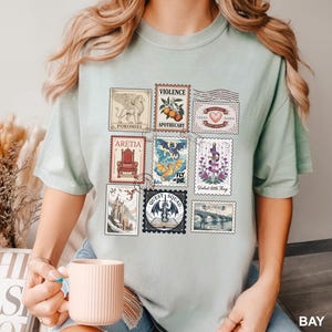 May include: A light green t-shirt featuring a collection of postage stamp designs. The stamps depict various images, including a griffin, a dragon, a heart, and a castle. Text on the stamps includes "POROMIEL", "VIOLENCE", "ARETIA", and "QUEST SQUAD".