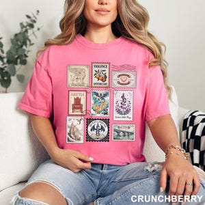 May include: A pink t-shirt featuring a collection of postage stamp designs. The stamps showcase various illustrations, including a griffin, a dragon, and a heart. The text "CRUNCHBERRY" is printed at the bottom of the shirt.