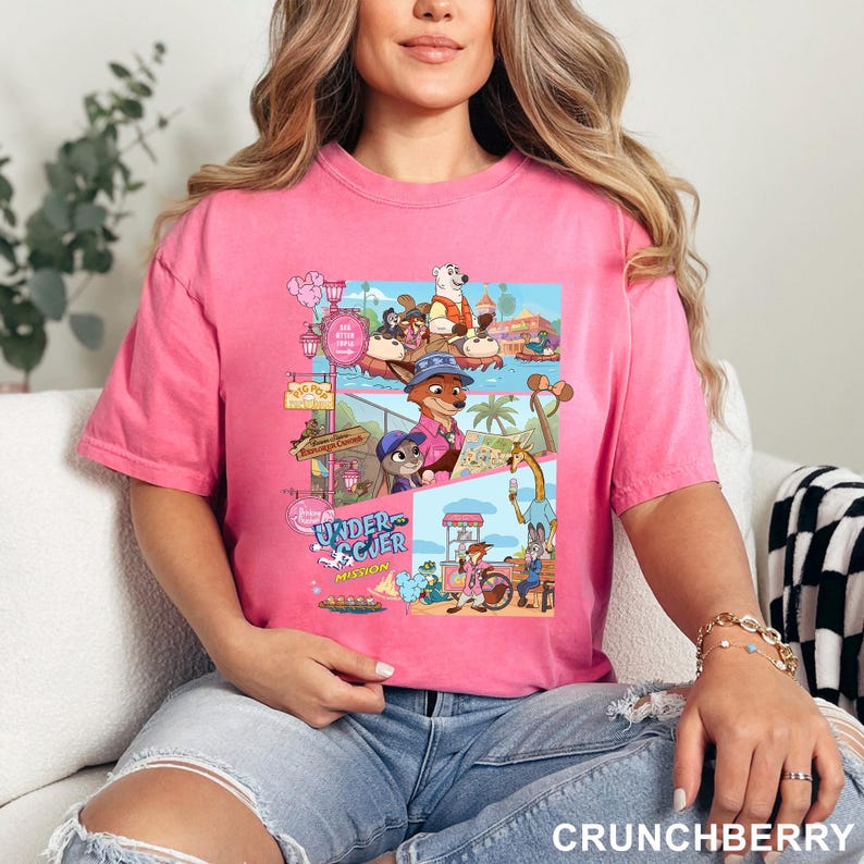 May include: A bright pink short-sleeved t-shirt featuring a multi-panel graphic print of various animated animal characters in a theme park setting. The graphic includes text that reads Undercover Mission and depicts scenes with a fox, rabbit, and giraffe.