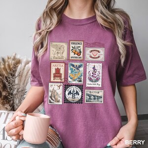 May include: Berry-colored t-shirt featuring a collection of vintage-style postage stamps. The stamps depict various illustrations, including a griffin, a dragon, and a bridge. Text on the stamps includes "VIOLENCE", "APOTHECARY", and "QUEST SQUAD".