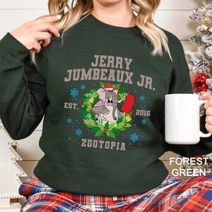 Pu&ograve; includere: Felpa verde foresta con grafica di Jerry Jumbeaux Jr. di Zootopia, con cappello di Babbo Natale e ghiacciolo rosso. Il testo "JERRY JUMBEAUX JR. EST. 2016 ZOOTOPIA" &egrave; stampato sul davanti.