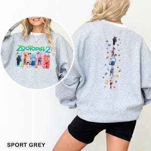 K&ouml;nnte beinhalten: Sportgraues Sweatshirt mit dem Text "ZOOTOPIA 2" und farbenfrohen Charaktergrafiken auf der Vorderseite. Die R&uuml;ckseite des Sweatshirts hat vertikale Charaktergrafiken. Das Sweatshirt ist aus einem weichen Material.