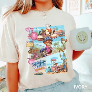 May include: An ivory t-shirt featuring a colorful graphic print of animated characters from Zootopia in a theme park setting. The design includes various scenes, park signage, and the text Undercover Mission. A light-colored mug with a smiley face is visible.