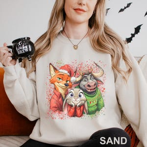 May include: A sand-colored sweatshirt featuring a colorful graphic of three cartoon characters: a fox in a Santa hat, a bunny, and a bull. The graphic is decorated with red and green paint splatters. The word "SAND" is printed at the bottom.