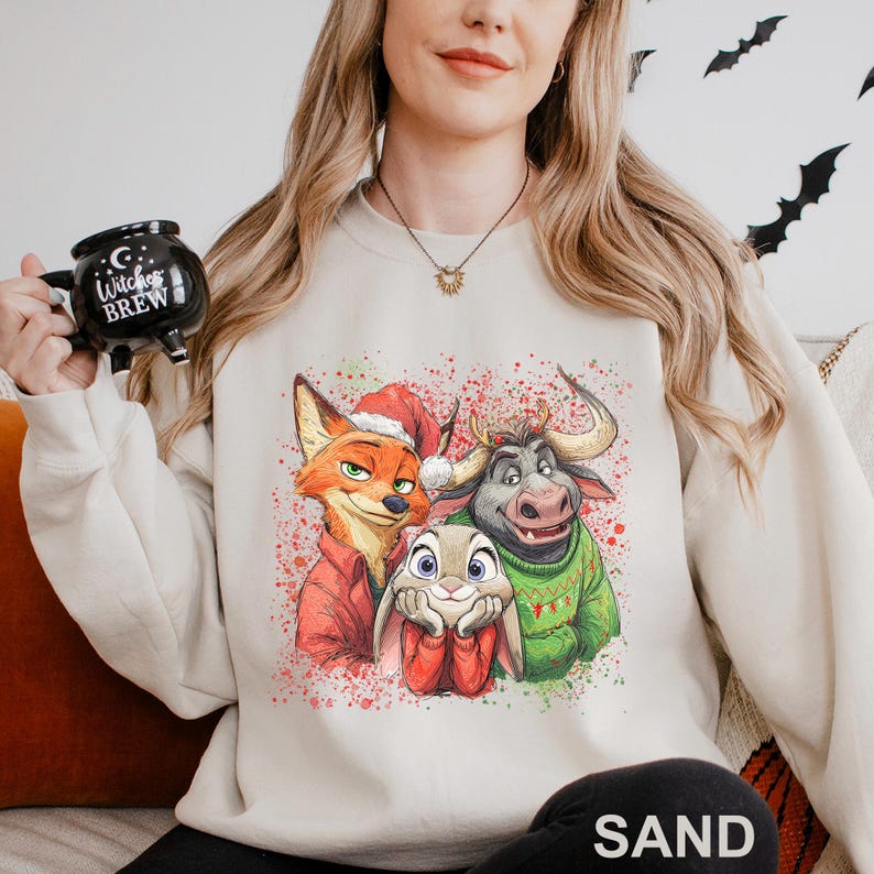 May include: A sand-colored sweatshirt featuring a colorful graphic of three cartoon characters: a fox in a Santa hat, a bunny, and a bull. The graphic is decorated with red and green paint splatters. The word "SAND" is printed at the bottom.