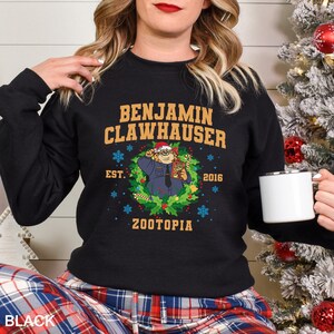 Pu&ograve; includere: Felpa girocollo nera con la scritta "BENJAMIN CLAWHAUSER" in oro. Il design include un personaggio dei cartoni animati in una ghirlanda con la scritta "ZOOTOPIA" e l'anno "2016". La felpa &egrave; indossata con pantaloni a quadri.