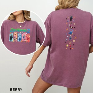 Op de afbeelding: Bessenkleurig T-shirt met een kleurrijke afbeelding van "Zootopia 2" op de voorkant en een verticaal ontwerp op de achterkant. De afbeelding op de voorkant bevat de tekst "Zootopia 2". Het ontwerp op de achterkant toont verschillende karakters en symbolen.
