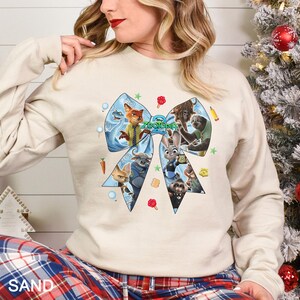 May include: A sand-colored sweatshirt featuring a large blue bow design with characters from the movie "Zootopia". The bow contains images of a fox, a bunny, a sloth, and other characters. The word "Zootopia" is in the center of the bow.