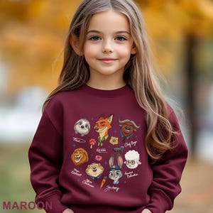 K&ouml;nnte beinhalten: Ein weinrotes Sweatshirt mit Zeichentrickfiguren aus dem Film Zoomania. Das Design umfasst Bilder von Flash, Nick Wilde, Chief Bogo, Judy Hopps und anderen. Das Wort "MAROON" ist unten aufgedruckt.