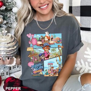 May include: A charcoal gray short-sleeve t-shirt featuring a multi-panel graphic print of animated animal characters in a theme park setting. The design includes the text "UNDERCOVER MISSION" and various scenes of foxes, rabbits, and other animals.