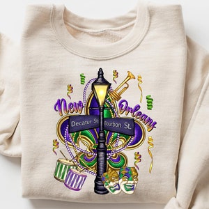 May include: A beige sweatshirt with a graphic print of a New Orleans street sign with a fleur-de-lis, a street lamp, a trumpet, and Mardi Gras masks.