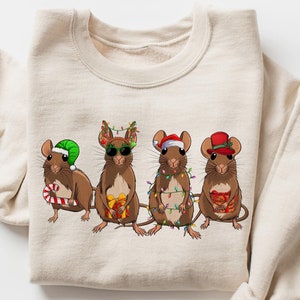 May include: A beige sweatshirt with a Christmas-themed design featuring four brown mice wearing festive hats and holding gifts. The mice are decorated with Christmas lights.