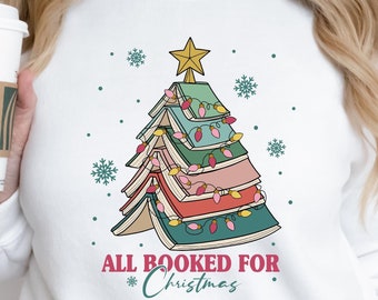 May include: White sweatshirt with a Christmas tree made of stacked books with lights. The text "All Booked for Christmas" is below the tree.