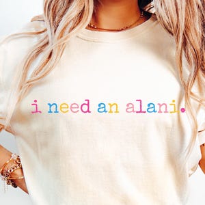 May include: Cream-colored t-shirt with the text "i need an alani." in rainbow colors. The shirt is paired with light blue denim jeans. The overall style is casual and relaxed.