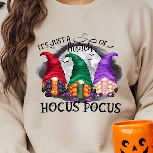 May include: A beige sweatshirt with the text 'It's just a bunch of Hocus Pocus' printed on it. The text is surrounded by three gnomes wearing hats in red, green, and purple. The gnomes are standing in front of a full moon.