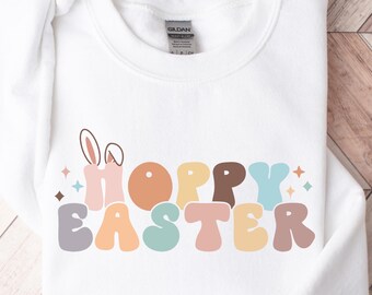 May include: White sweatshirt with a colorful "Hoppy Easter" graphic featuring a bunny with a carrot. The letters are in a retro style with pastel colors.