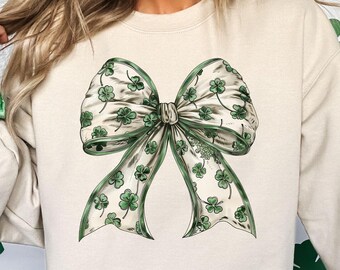May include: A cream-colored sweatshirt with a green shamrock bow design. The bow is made of a white ribbon with green shamrocks printed on it.