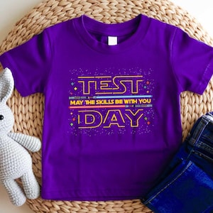 May include: A purple t-shirt with the words "TEST DAY" in yellow, with the phrase "MAY THE SKILLS BE WITH YOU" in white. The design includes blue and red lightsaber-like elements. The shirt is paired with blue jeans.