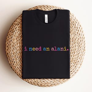 May include: Black crew neck t-shirt with the text "i need an alani." in rainbow colors. The shirt is displayed on a round, woven, beige surface. The shirt has a tag on the inside of the collar.