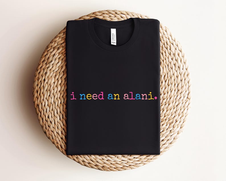 May include: Black crew neck t-shirt with the text "i need an alani." in rainbow colors. The shirt is displayed on a round, woven, beige surface. The shirt has a tag on the inside of the collar.