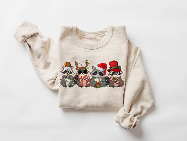 May include: A beige sweatshirt with a Christmas-themed design featuring four raccoons wearing festive hats and holding Christmas lights and candy canes.