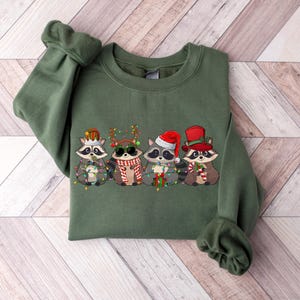 May include: A green Christmas sweatshirt with a design of five raccoons wearing Santa hats and holding Christmas lights. The raccoons are wearing different hats and holding different Christmas items.