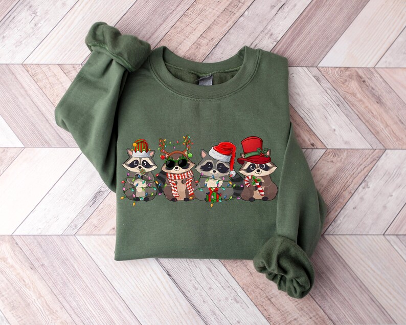 May include: A green Christmas sweatshirt with a design of five raccoons wearing Santa hats and holding Christmas lights. The raccoons are wearing different hats and holding different Christmas items.