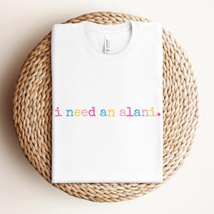 May include: White t-shirt with the text "i need an alani." in rainbow colors. The shirt is folded and placed on a round, woven, light brown surface.