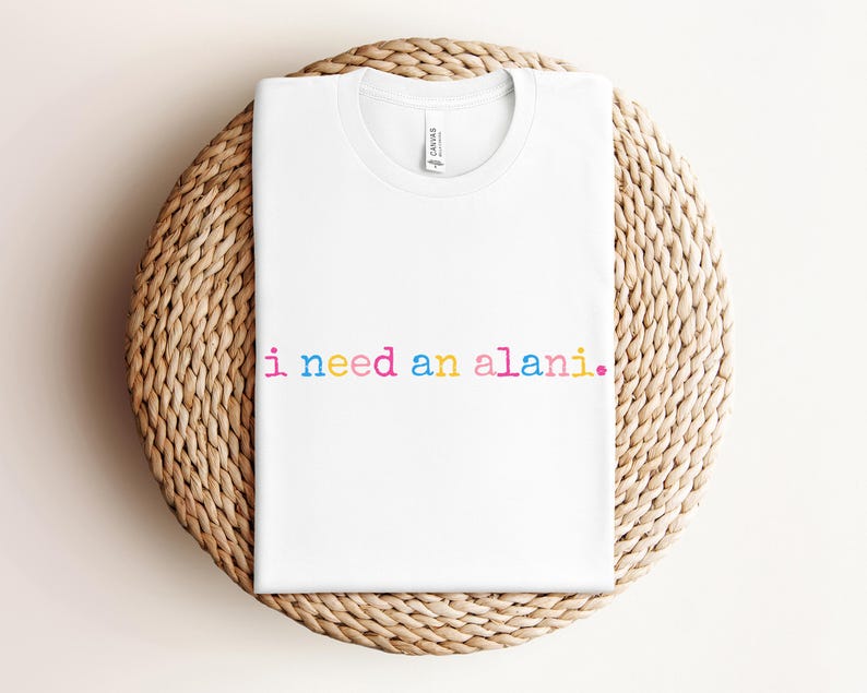 May include: White t-shirt with the text "i need an alani." in rainbow colors. The shirt is folded and placed on a round, woven, light brown surface.