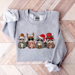 May include: Gray sweatshirt with a Christmas-themed design featuring four raccoons wearing Santa hats and holding candy canes and lights.