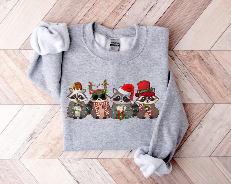 May include: Gray sweatshirt with a Christmas-themed design featuring four raccoons wearing Santa hats and holding candy canes and lights.