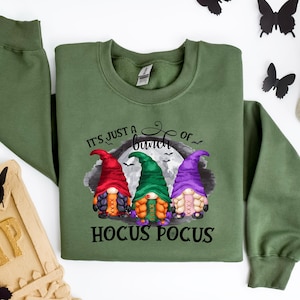 以下が含まれることがあります： 「It's just a bunch of Hocus Pocus」の文字と3人のノームのキャラクターが描かれたオリーブグリーンのスウェットシャツ。ノームは赤、緑、紫の帽子をかぶっています。黒い蝶と骸骨が背景にあります。