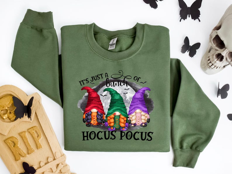 以下が含まれることがあります： 「It's just a bunch of Hocus Pocus」の文字と3人のノームのキャラクターが描かれたオリーブグリーンのスウェットシャツ。ノームは赤、緑、紫の帽子をかぶっています。黒い蝶と骸骨が背景にあります。
