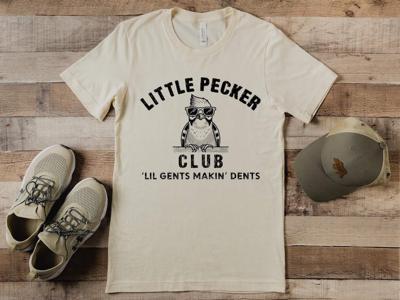 May include: A beige t-shirt with a black and white illustration of a woodpecker wearing sunglasses. The text "Little Pecker Club 'Lil Gents Makin' Dents" is printed on the shirt.