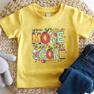 May include: A yellow t-shirt with the text "you are MORE than a test SCORE" in colorful block letters. The design includes pencils, rulers, and stars. The shirt is paired with blue denim jeans.