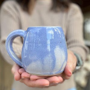 May include: A handmade, ceramic mug in a soft periwinkle blue with white accents. The mug has a rounded body, a large handle, and a slightly textured surface. The mug is held in both hands, suggesting a cozy, comforting feel. The mug is perfect for coffee or tea.