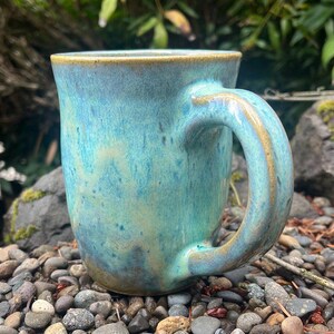 May include: A ceramic mug with a glossy glaze featuring a blend of turquoise, teal, and soft blue tones. The mug has a rounded body, a thick curved handle, and a smooth rim with subtle brown undertones visible along the edges and base.