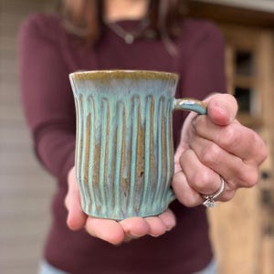 May include: A handmade ceramic mug with a vertical ribbed design. The mug features a turquoise glaze with brown accents and a brown rim. The mug is held in both hands, showcasing its unique shape and color.