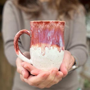 May include: A handmade ceramic mug with a unique glaze. The top half of the mug is a deep red with a dripped effect, transitioning to a speckled white base. The handle is red, complementing the mug's design. A perfect addition to any kitchen.