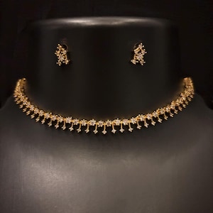 May include: A gold-coloured necklace and matching earrings set. The necklace is a delicate choker style with small, dangling accents. The earrings are small clusters of stones. The jewellery is displayed on a black mannequin.