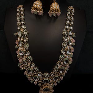 Op de afbeelding: Een goudkleurige ketting- en oorbellenset. De ketting heeft heldere, ovaalvormige stenen en kleinere groene en roze accenten. De oorbellen zijn klokvormig met vergelijkbare stenen en roze kralen. De sieraden worden op een zwarte standaard gepresenteerd.