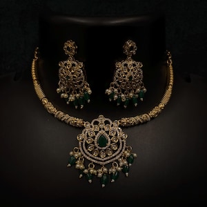 May include: Gold-coloured necklace and earring set. The necklace has a pendant with a central green stone, surrounded by clear stones and dangling white pearls and green beads. The earrings mirror the pendant design, with dangling pearls and green beads.